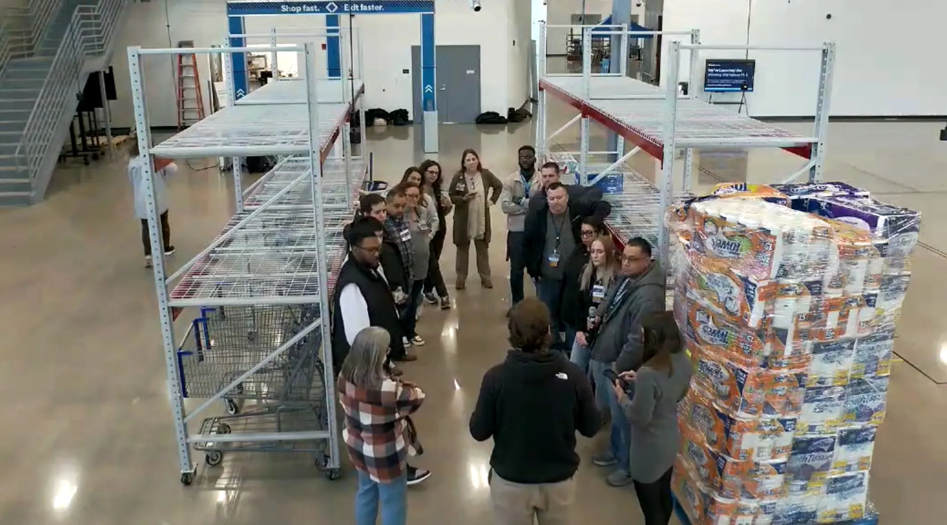 Overhead view of a testing session at The Clubhouse: associates and managers gathered beside a wrapped pallet of paper towels and empty steel shelving.